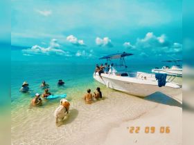 A lively group enjoys a beach day with a boat anchored nearby, swimming and relaxing in the clear waters — perfect for summer fun and memorable outings.
