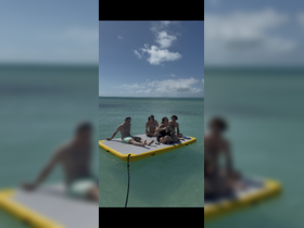 A lively group enjoys a sunny day on a floating mat in clear turquoise waters, perfect for relaxation and fun in the sun.
