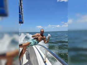 A man enjoys a thrilling moment on a boat, playfully posing with a hat against a backdrop of clear blue skies and sparkling waters — perfect for capturing the joy of a day on the water.