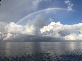 A stunning view of a vibrant rainbow arching over calm ocean waters, framed by dramatic clouds — a picturesque moment capturing the beauty of nature at sea.