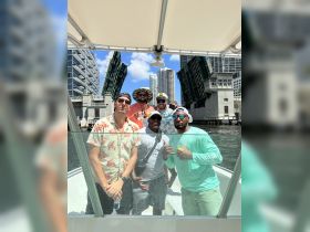A lively group of friends celebrates on a boat as they pass under a raised bridge, enjoying drinks and the sunny weather — perfect for a fun day out on the water.