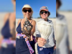Two friends proudly display their shell finds on the beach, capturing a fun moment during their coastal adventure.