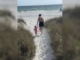 A mother and her young daughter stroll hand-in-hand along a sandy path leading to the beach, capturing a heartwarming family moment by the ocean.