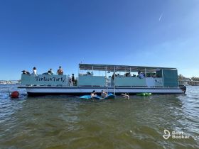 A lively group enjoys a fun-filled day on a pontoon party boat, with guests swimming and relaxing in the water under a clear blue sky.