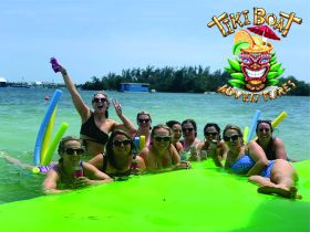 A lively group of friends enjoys a fun day on the water with drinks and pool noodles, celebrating their Tiki Boat adventure in the sun.