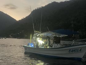 Two friends enjoy an evening fishing trip aboard the boat 'Camila,' surrounded by stunning mountain scenery as the sun sets.