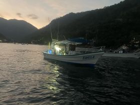 The fishing boat 'Camila' is illuminated at dusk, with people onboard enjoying an evening on the water, showcasing a serene atmosphere perfect for fishing adventures.