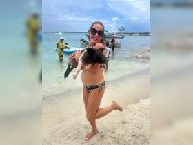 A woman joyfully holds a pig at the beach, showcasing a unique human-animal interaction in a tropical setting.