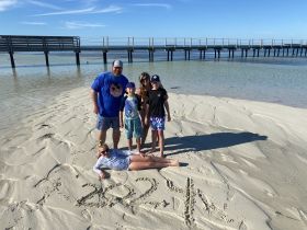 A joyful family poses on the beach, with a pier in the background and playful sand drawings — a perfect moment capturing fun in the sun.