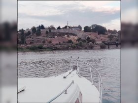 A serene view of the bow of a boat cruising along a river, with a picturesque landscape featuring a historic fortification in the background.