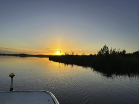 A serene sunset view over calm waters, with the silhouette of vegetation and the edge of a boat visible in the foreground — perfect for tranquil evening cruises.