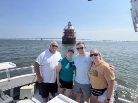 A cheerful group poses for a photo on their boat with a historic lighthouse in the background, capturing a memorable moment during their water adventure.