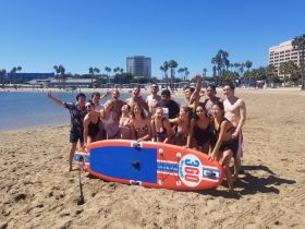 A lively group of friends poses for a fun photo on the beach with a paddleboard, capturing the essence of summer celebrations and beach activities.