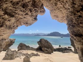A stunning view from a rocky archway overlooking a serene beach and turquoise waters, with a boat gently anchored in the distance — a perfect representation of coastal beauty.