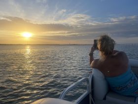 A woman captures the stunning sunset from the deck of a pontoon boat, creating a perfect moment of relaxation on the water.