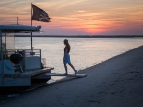 A woman walks towards a boat adorned with a pirate flag at sunset, creating a picturesque scene perfect for evening adventures on the water.