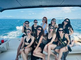 A lively group of women celebrates aboard a yacht, showcasing a fun bachelorette party atmosphere with stylish outfits and smiles against a stunning ocean backdrop.