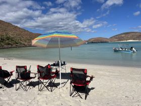 A serene beach scene featuring colorful chairs and an umbrella, with a boat anchored in the calm waters — perfect for a relaxing day by the shore.