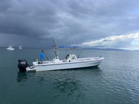 A man stands on a fishing boat in calm waters, surrounded by other vessels under a dramatic sky — a perfect setting for a fishing adventure.