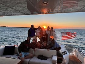 A joyful group of friends gathers on a yacht at sunset, capturing a memorable moment with the vibrant colors of the sky as a backdrop — perfect for celebrations and gatherings on the water.