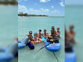 A fun group of friends enjoys a day at the beach, relaxing on inflatable floats and paddleboards in crystal-clear waters — perfect for summer adventures and celebrations.
