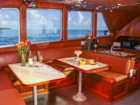 A cozy interior view of a boat's dining area, featuring elegant seating, a table set with wine glasses, and a beautiful floral arrangement, perfect for intimate meals while enjoying the ocean view.