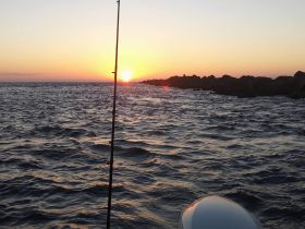 A serene sunset view over the water, with a fishing rod in the foreground, capturing the essence of a peaceful fishing trip at dusk.