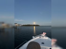 A serene view from the front of a boat, capturing the reflection of the moonlight on the water, with a distant island and docked boats in the background.