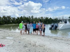 A cheerful group gathers on the beach near a pontoon boat, enjoying a fun day at the sandbar — perfect for celebrations and group outings.