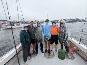 A group of friends poses for a photo on a fishing boat, dressed in rain gear, ready for an adventurous day on the water despite the overcast weather.