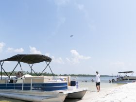 Guests enjoy a sunny day at the beach with pontoon boats nearby, while an airshow unfolds overhead — a perfect blend of relaxation and excitement on the water.