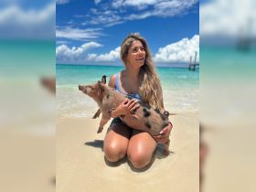 A woman enjoys a playful moment with a pig on the beach, showcasing a unique wildlife interaction in a tropical paradise.