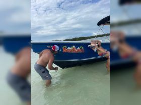 A couple enjoys a playful moment by their boat named 'Main Squeeze' in shallow waters, capturing the essence of fun and relaxation on the beach.