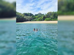 A couple enjoys a refreshing swim in crystal-clear waters, surrounded by lush greenery and a serene beach backdrop — perfect for tropical adventures.