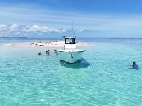A group enjoys a fun day at a sandbar, swimming in crystal-clear turquoise waters with a boat anchored nearby — a perfect tropical getaway.