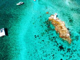 A stunning aerial view of boats anchored near a rocky island, with people swimming in the vibrant turquoise waters — a perfect scene for adventure and relaxation.