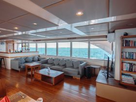 A spacious and inviting interior lounge area of a boat, featuring comfortable seating and large windows offering stunning views of the ocean — perfect for relaxation during a cruise.
