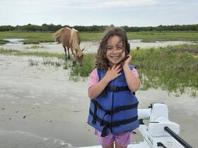 A joyful child in a life jacket smiles at the camera while a wild horse grazes in the background, capturing a unique moment of human-animal interaction on the beach.