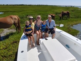 A joyful family poses on their boat with wild horses grazing in the background, capturing a memorable outdoor adventure.