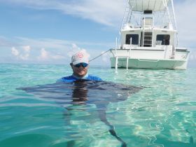 A man interacts with a stingray in the clear turquoise waters, showcasing a unique wildlife experience while a boat is anchored nearby.