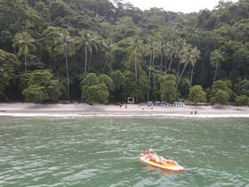 A couple enjoys paddleboarding in a serene tropical setting, with a beautiful beach and lush greenery in the background — perfect for a romantic getaway.