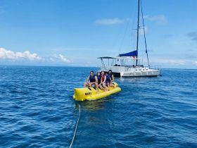 A group of friends enjoys a thrilling ride on a banana boat, with a beautiful sailing catamaran in the background — perfect for adventurous water activities.