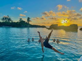 A group of friends enjoys a lively sunset swim, with one person jumping into the water, creating a perfect moment of fun and celebration against a stunning backdrop.