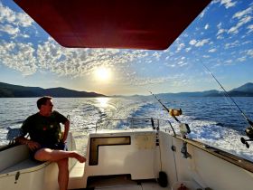A man relaxes on the deck of a boat during a fishing trip at sunset, with stunning views of the mountains and water creating a perfect backdrop for a memorable experience.