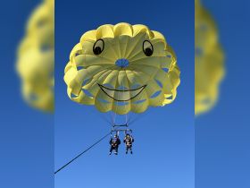 Two adventurers enjoy a thrilling parasailing experience under a bright yellow smiley face parachute, soaring high against a clear blue sky.
