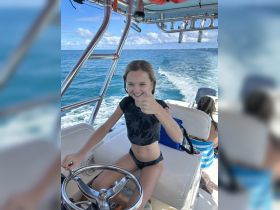 A young girl enthusiastically gives a thumbs up while steering a boat, capturing the joy of on-water adventures.