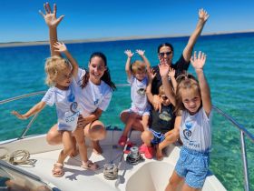 A joyful group of children and adults celebrate on a boat, waving their hands in excitement against a stunning turquoise backdrop — perfect for family outings and fun on the water.