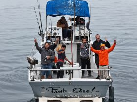 A lively group of friends celebrates their fishing trip aboard 'The Fresh One!' with smiles and peace signs, showcasing the joy of a day on the water.