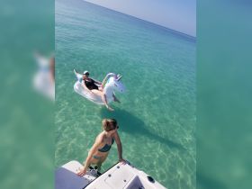 Two friends enjoy a sunny day in clear waters, one relaxing on a unicorn float while the other wades near the boat — a perfect moment for summer fun and relaxation.