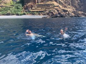 A couple enjoys a refreshing swim in crystal-clear waters, waving at the camera amidst stunning coastal scenery — a perfect moment of fun and adventure.